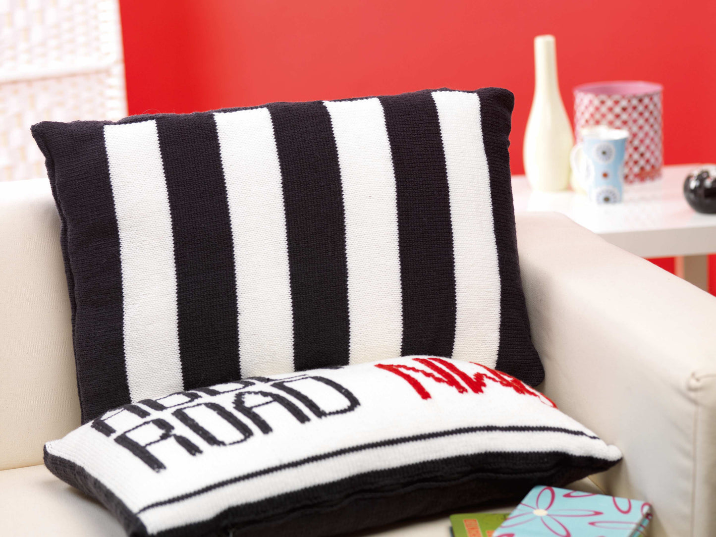 Image shows two knitted cushions one showing the abbey road street sign and behind it a second black and white striped cushion looking like the zebra crossing from the beatles music album knitting pattern by jane burns