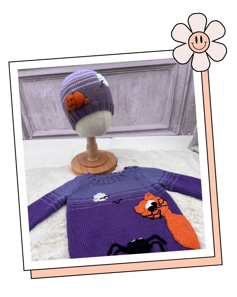 A purple knit baby hat with a pumpkin patch and a matching purple knit long-sleeve sweater featuring a fox, spider, and moon design, displayed on a mannequin stand and fabric surface.