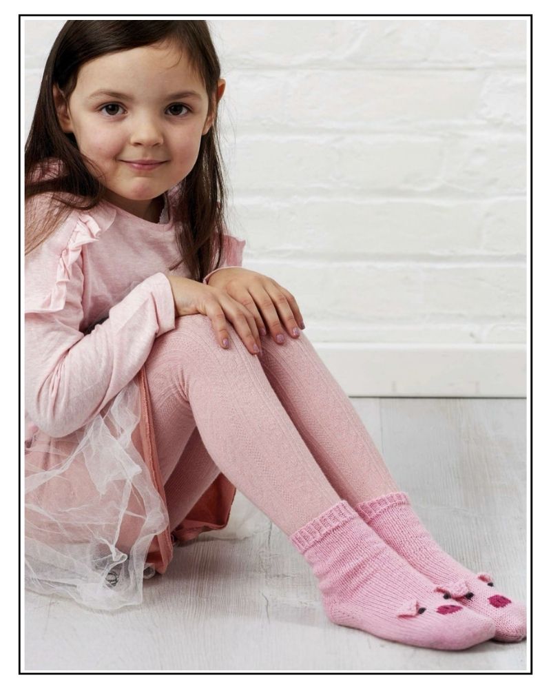 Pig socks knitting pattern, young girl sitting by a white wall wearing pink hand knitted socks with pig faces
