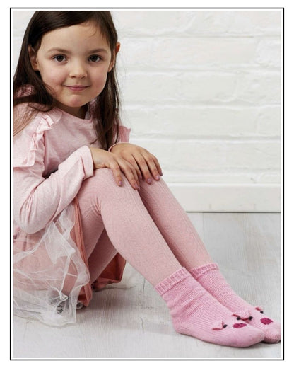 Pig socks knitting pattern, young girl sitting by a white wall wearing pink hand knitted socks with pig faces