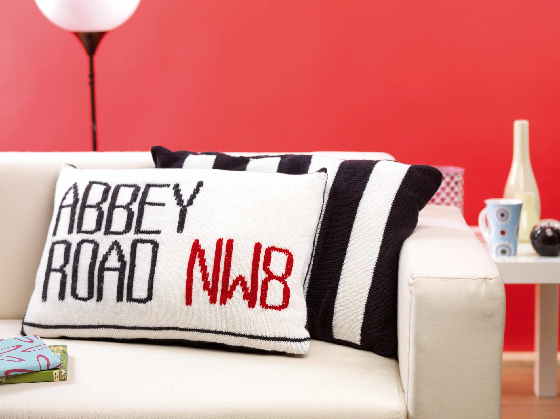 Image shows two knitted cushions one showing the abbey road street sign and behind it a second black and white striped cushion looking like the zebra crossing from the beatles music album knitting pattern by jane burns