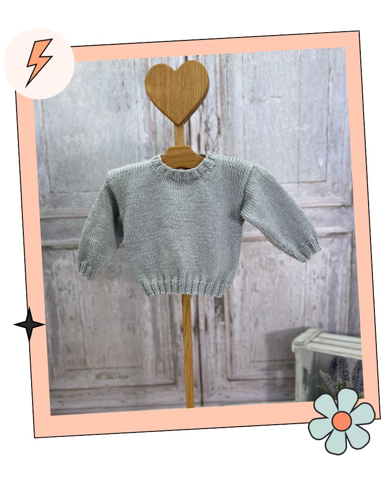 A light gray knit baby sweater displayed on a wooden hanger with a heart-shaped top, set against a rustic gray wooden door background with a peach-colored border and decorative graphic elements. The Blank Canvas Baby Sweater, design your own baby jumper using the motif library by jane burns