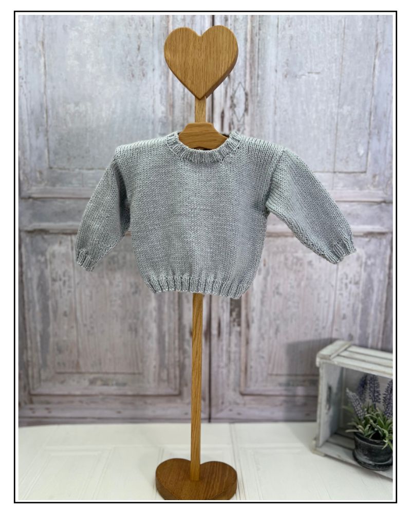 A light gray knit baby sweater displayed on a wooden hanger with a heart-shaped top and stand, set against a rustic distressed gray wooden door background. The Blank Canvas Baby Sweater, design your own baby jumper using the motif library by jane burns