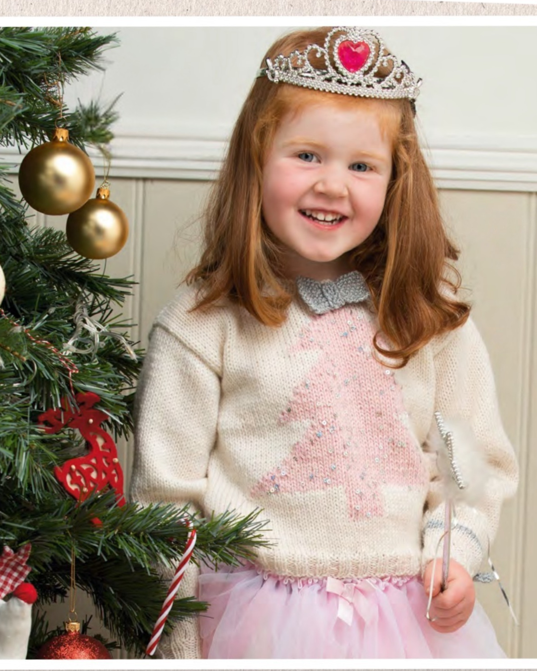 A young girl with red hair smiling next to a decorated Christmas tree, wearing a cream knit sweater with a pink sequined Christmas tree, a silver tiara, and a pink tutu skirt.