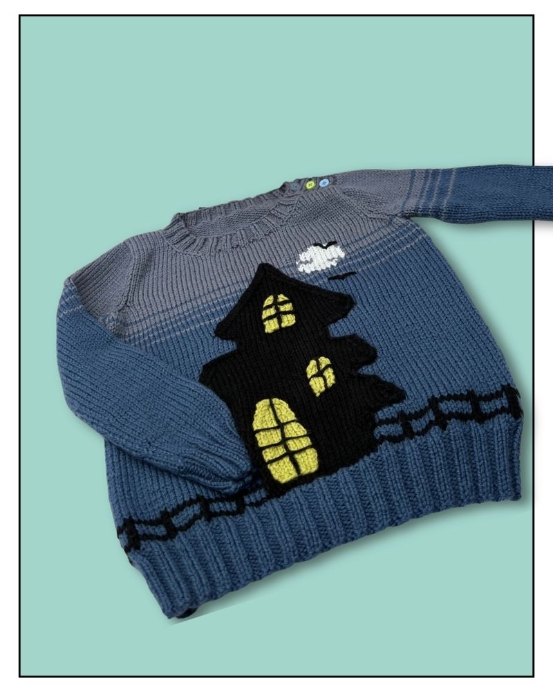 A blue and gray striped knit sweater featuring a large black haunted house with yellow glowing windows on the front, displayed on a solid mint green background.