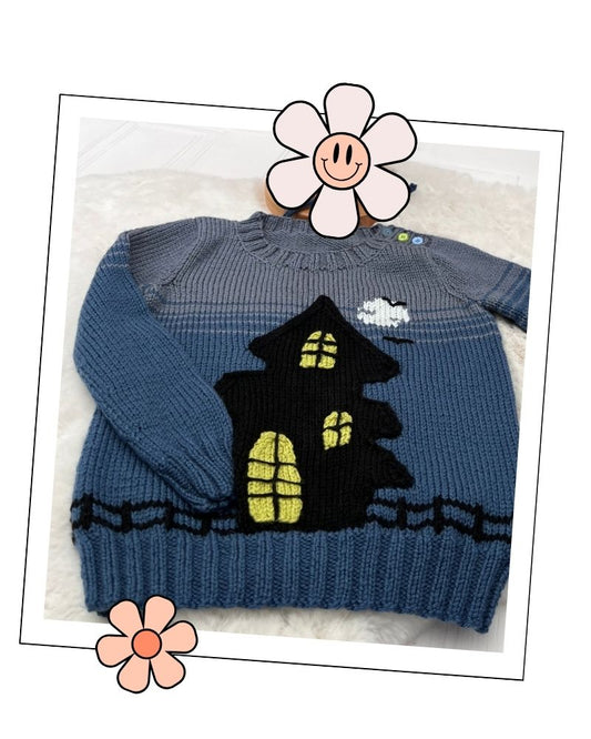 A blue knit baby sweater featuring a black haunted house with yellow glowing windows and a small white cloud motif, presented on a white textured background with decorative flower graphics.