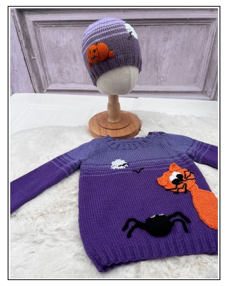 A purple knit sweater and matching beanie displayed on a mannequin stand and surface. The sweater has orange, black, and white Halloween-themed appliqués including a fox, spider, moon, and stars. Halloween hats collection knitting patterns by jane burns