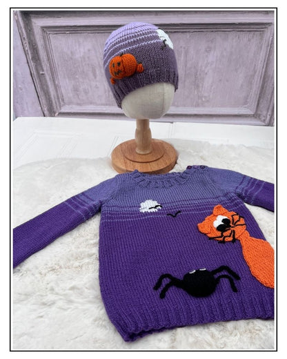 A purple knit sweater and matching beanie displayed on a mannequin stand and surface. The sweater has orange, black, and white Halloween-themed appliqués including a fox, spider, moon, and stars. Halloween hats collection knitting patterns by jane burns