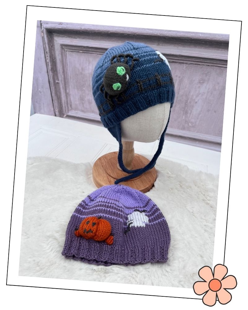 Two knitted Halloween-themed baby hats displayed on a mannequin head and a wooden stand. The top hat is dark blue with a black spider and green eyes; the bottom hat is purple with a purple-striped ghost, white cloud, and orange jack-o'-lantern.