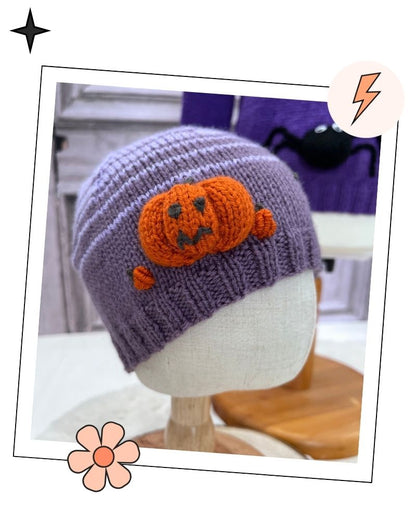 A purple knit beanie hat with an orange knitted jack-o'-lantern decoration on the front, displayed on a white mannequin head. The photo is styled inside a white border with decorative star, flower, and lightning bolt graphics.
