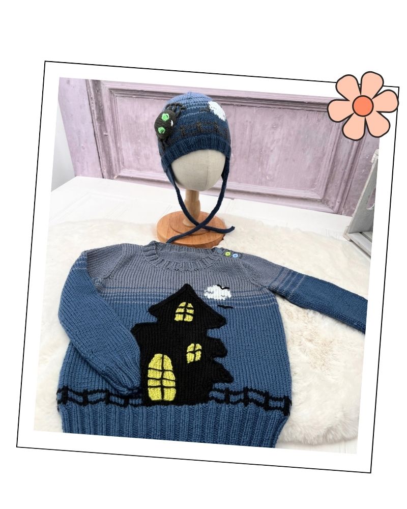 A blue and gray knit sweater with a black haunted house design and yellow window details, displayed on a white faux-fur surface with a matching blue knit hat on a mannequin stand in the background.