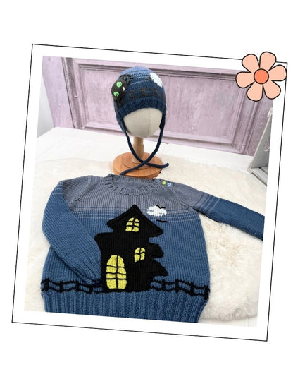 A blue and gray knit sweater with a black haunted house design and yellow window details, displayed on a white faux-fur surface with a matching blue knit hat on a mannequin stand in the background.