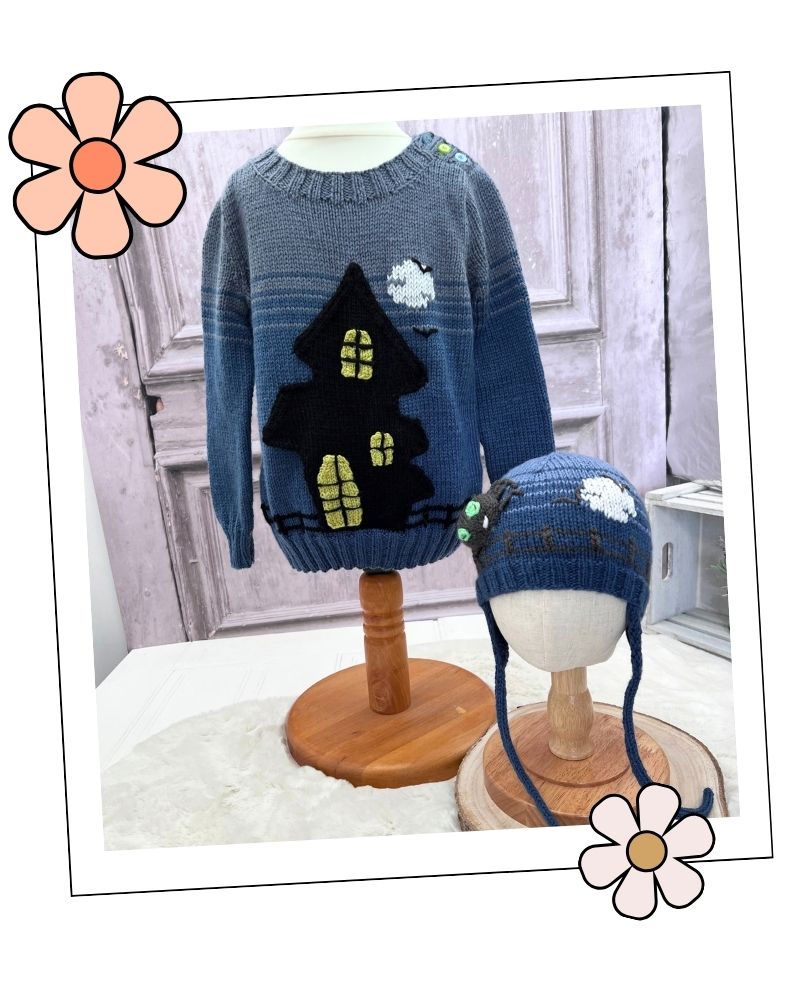 A blue knit sweater and matching blue knit hat displayed on mannequin stands in an indoor styled setting. The sweater features a large black house with yellow window details and a small white cloud motif. Halloween hats collection knitting patterns addition to the ghost house jumper shown