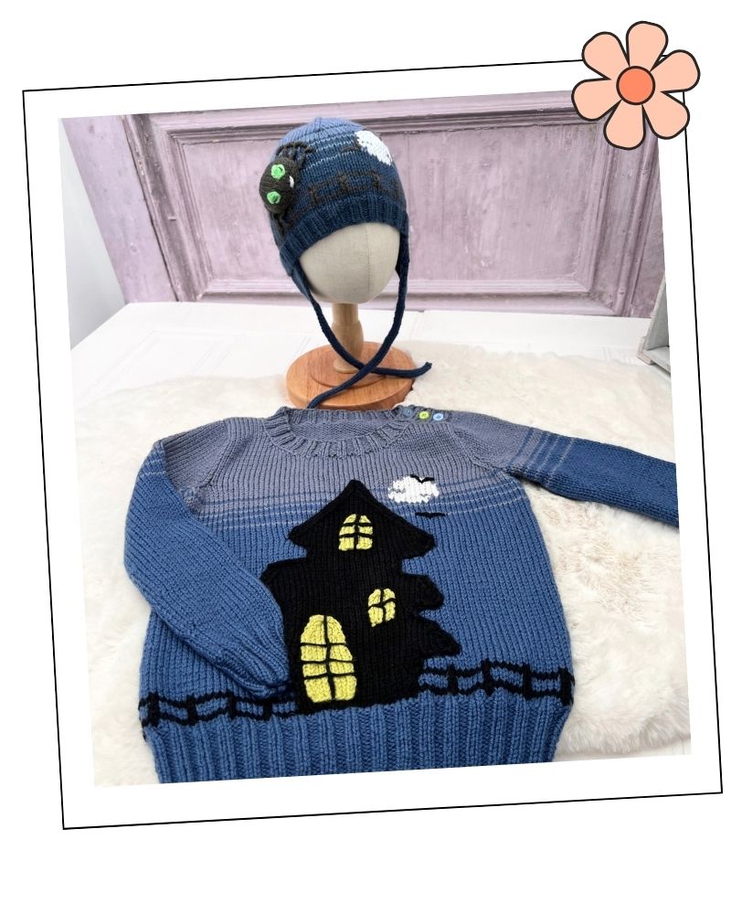 Mix and Match Halloween Hats Knitting Pattern Collection by Jane BurnsA blue knit children's sweater with a black haunted house and yellow windows, displayed on a white faux-fur surface with a matching blue knit hat on a mannequin head in the background.
