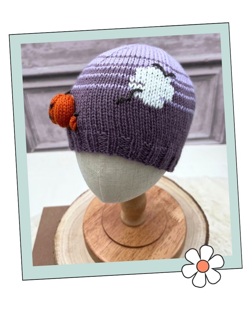 Pumpkin Hat with full moon motif from halloween hats collection knitting pattern by Jane Burns.
A knitted beanie hat in lavender purple with a white cloud motif and an orange cloud accent, displayed on a mannequin head stand.