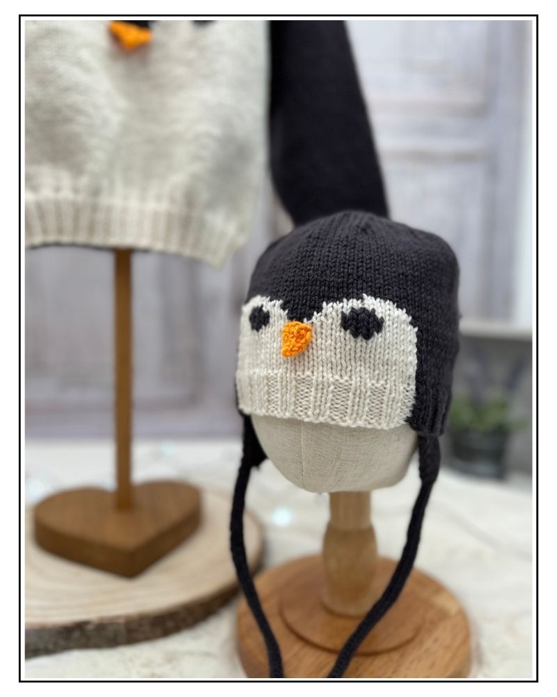 Penguin aviator hat with ear flaps and ties on a wooden hat stand, baby knitting pattern by Jane Burns