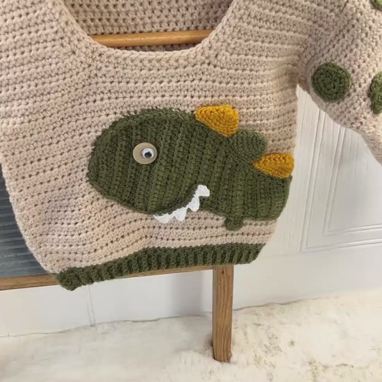 beige crochet sweater with moss green cuff, featuring a moss green dinosaur applique, mustard and moss green spikes run down the spine of the dinosaur. the dinosaur has white teeth and googly eye. the sleeves of the jumper has crochet spots sewn to them. dinotastic crochet sweater pattern for babies and toddlers by jane burns
