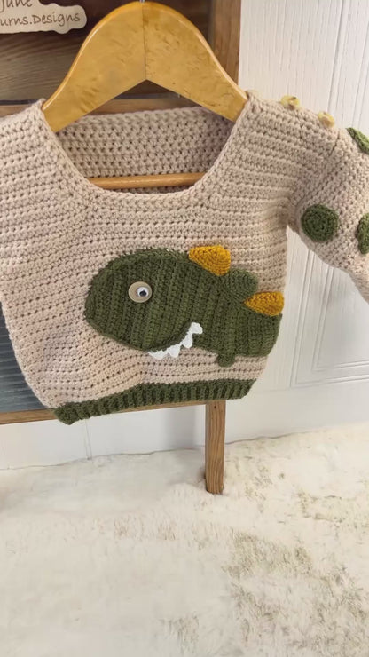 beige crochet sweater with moss green cuff, featuring a moss green dinosaur applique, mustard and moss green spikes run down the spine of the dinosaur. the dinosaur has white teeth and googly eye. the sleeves of the jumper has crochet spots sewn to them. dinotastic crochet sweater pattern for babies and toddlers by jane burns