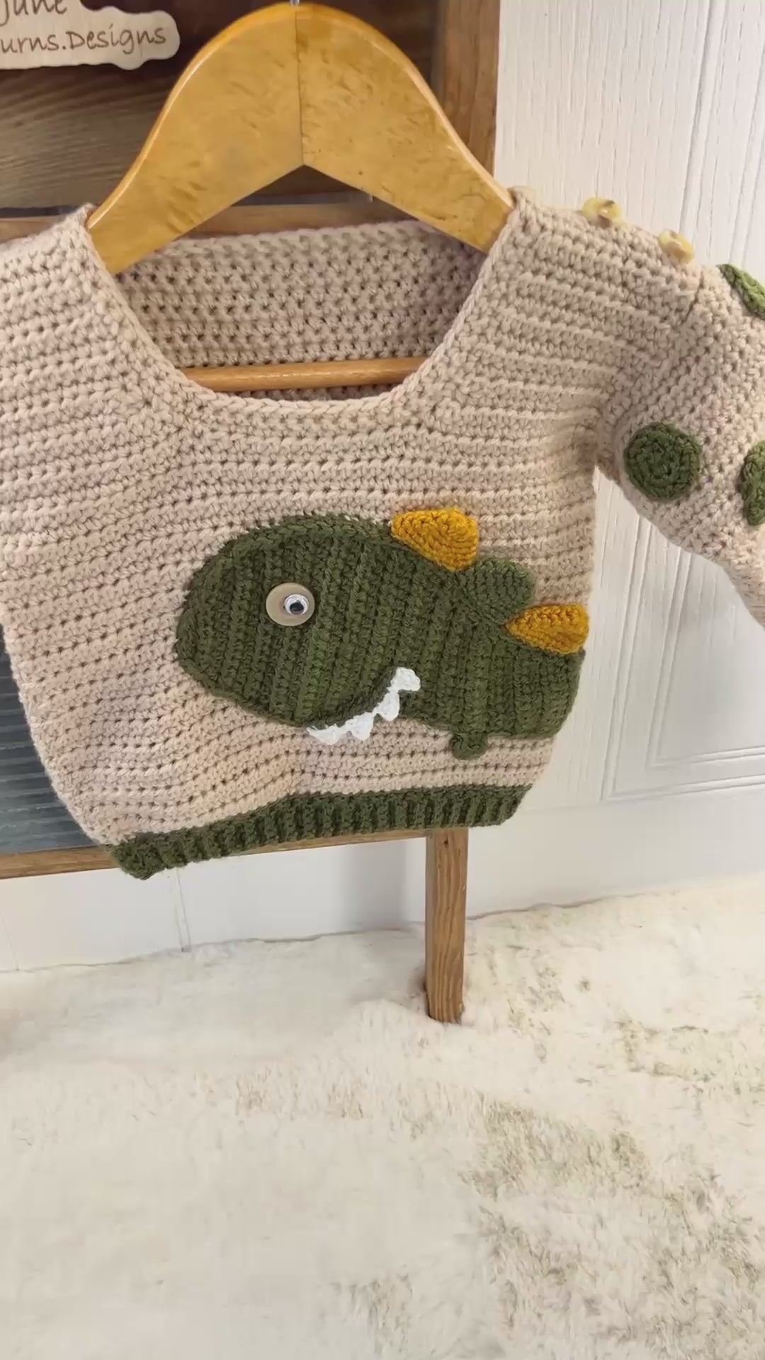 beige crochet sweater with moss green cuff, featuring a moss green dinosaur applique, mustard and moss green spikes run down the spine of the dinosaur. the dinosaur has white teeth and googly eye. the sleeves of the jumper has crochet spots sewn to them. dinotastic crochet sweater pattern for babies and toddlers by jane burns