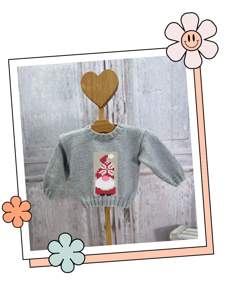 A light gray knit baby sweater with a red and white gnome graphic on the chest, displayed on a wooden hanger with a heart-shaped wooden hook against a rustic gray wood plank background. blank canvas baby jumper knitting pattern showing jumper with dinosaur motif and three other charts that you can use to create a unique jumper jane burns designs