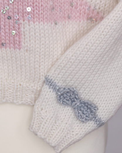 Close-up of a knitted sweater or garment on a mannequin, showing a cream knit body with a pink sequin band and a gray sequin bow detail on the sleeve cuff. sparkle christmas tree jumper for girls knitting pattern by jane burns