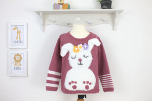 Pink kids sweater with white fluffy bunny motif, large floppy ears and applique flower garland on its head. Big bunny feet with pink toes. Knitting pattern by Jane Burns Designs